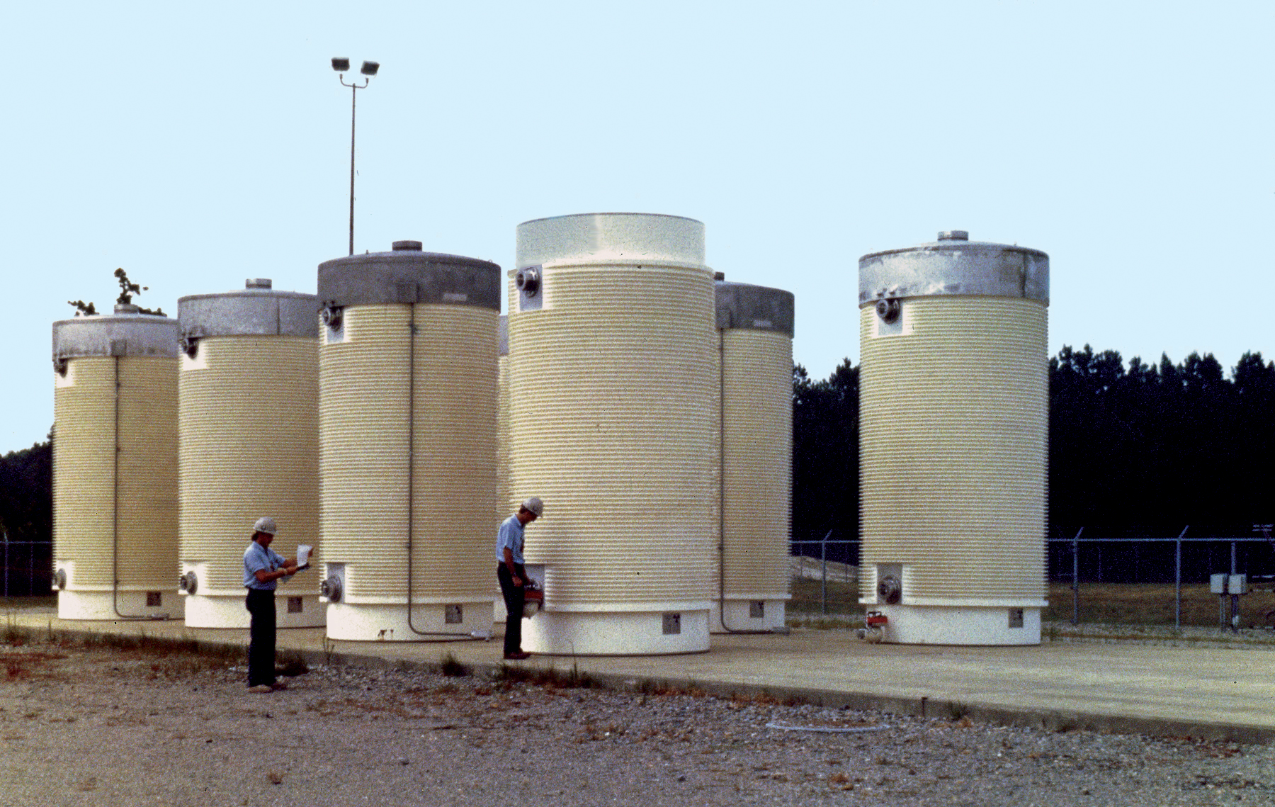 A row of vertical concrete-and-steel dry casks at an outdoor independent spent fuel storage installation, with two workers in hard hats nearby for scale.